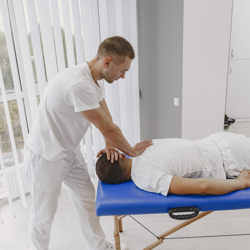 A person in a white shirt performs a neck and back adjustment on a person lying on a blue massage table in a bright room. A person in a white shirt performs a neck and back adjustment on a person lying on a blue massage table in a bright room