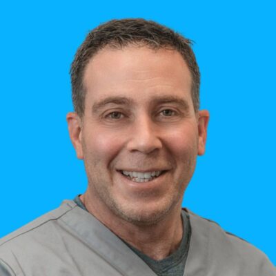 Dr. Gregg Greenblatt - Podiatrist A man with short brown hair and a grey shirt smiling in front of a solid blue background.
