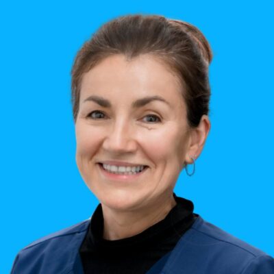 Woman with brown hair tied back, wearing a navy blue top and hoop earrings, smiling in front of a solid blue background. Woman with brown hair tied back, wearing a navy blue top and hoop earrings, smiling in front of a solid blue background.