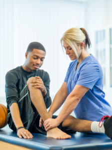 A physical therapist in scrubs assists a young man with a leg stretch on a padded table in a bright treatment room.