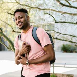 A young man wearing a pink shirt and backpack holds a book while smiling outdoors near a tree, just steps away from his favorite chiropractor in Lower Manhattan.