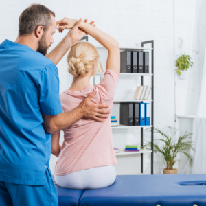 A healthcare professional in blue scrubs assists a woman seated on an exam table with a shoulder stretch in a Chiropractor Lower Manhattan medical office.