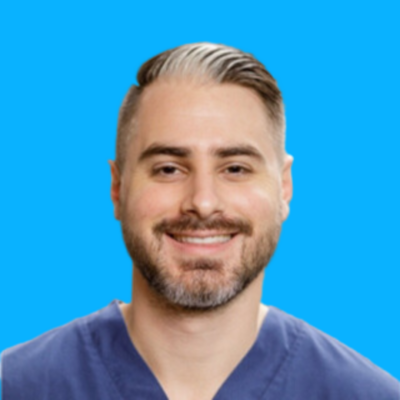 A man with short hair, a beard, and mustache is smiling while wearing blue medical scrubs against a solid blue background. A man with short hair, a beard, and mustache is smiling while wearing blue medical scrubs against a solid blue background.
