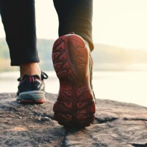 Close-up of a person in athletic shoes walking on a rocky surface near a body of water, with sunlight in the background—an inspiring scene for those seeking Physical Therapy In Manhattan to regain strength and mobility.