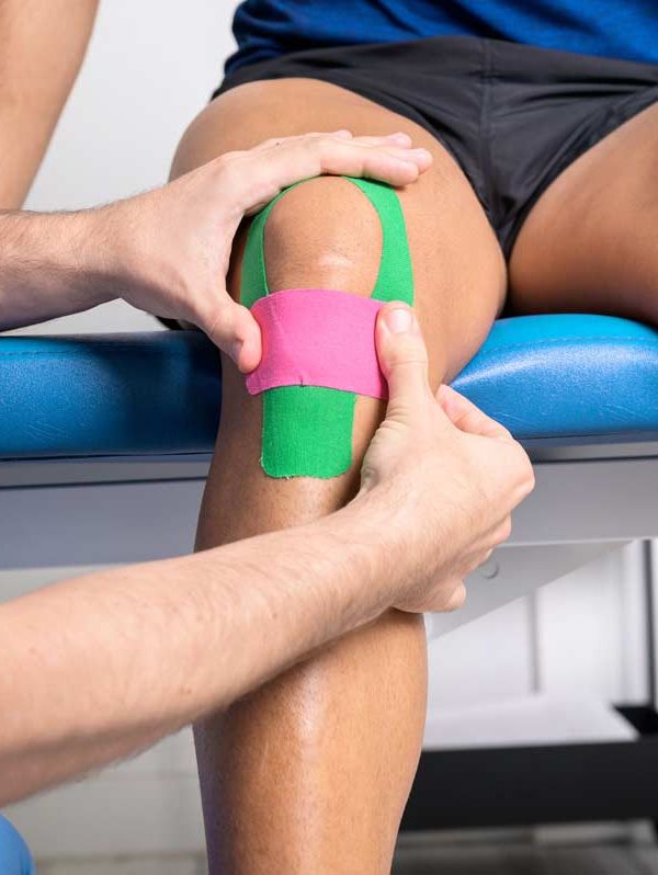 A medical professional applies green and pink kinesiology tape to a patient's knee while the patient sits on an exam table