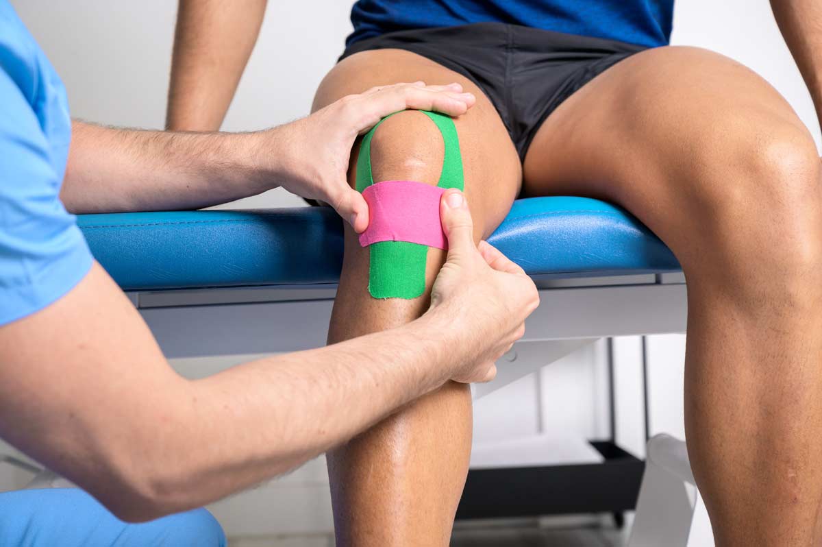 A medical professional applies green and pink kinesiology tape to a patient's knee while the patient sits on an exam table