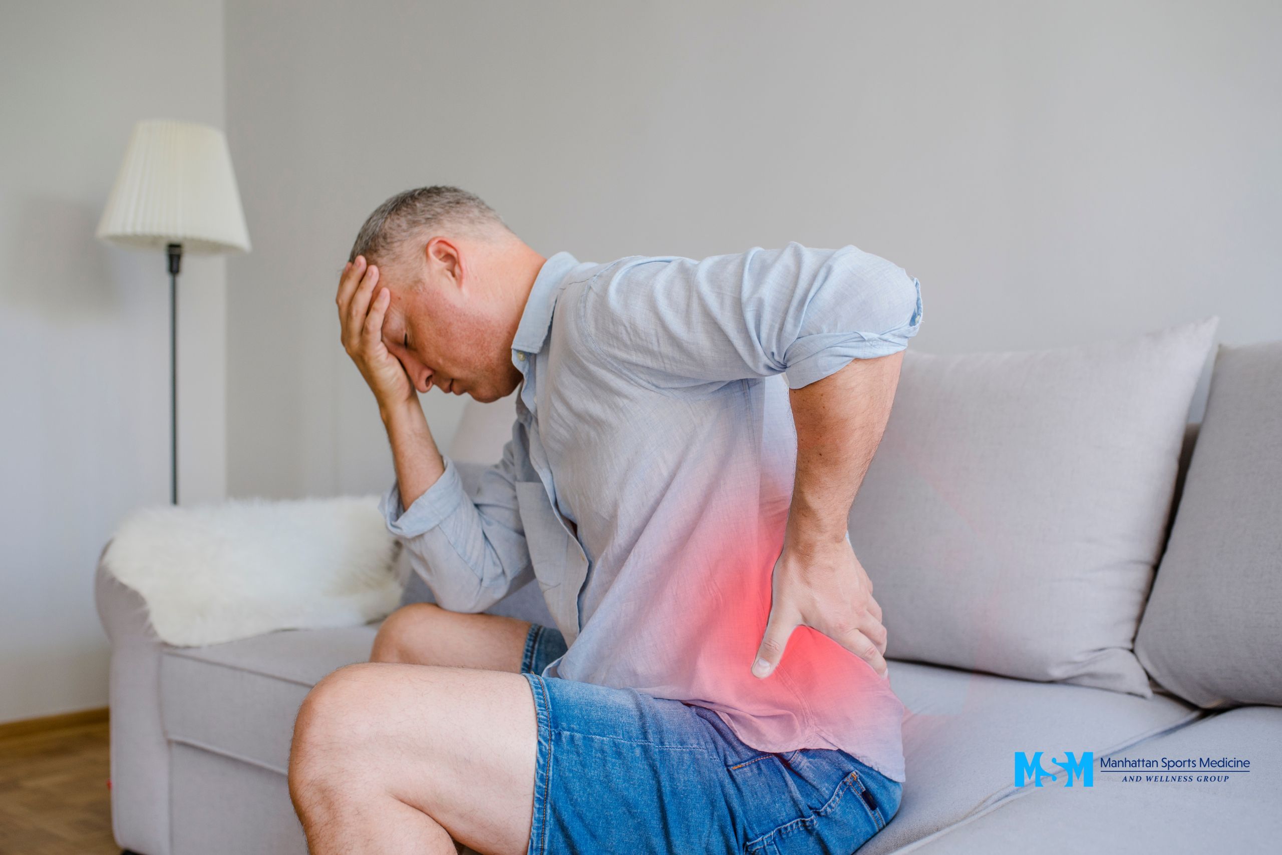 A man sitting on a couch holds his head with one hand and his lower back with the other, indicating pain. A man sitting on a couch holds his head with one hand and his lower back with the other, indicating pain