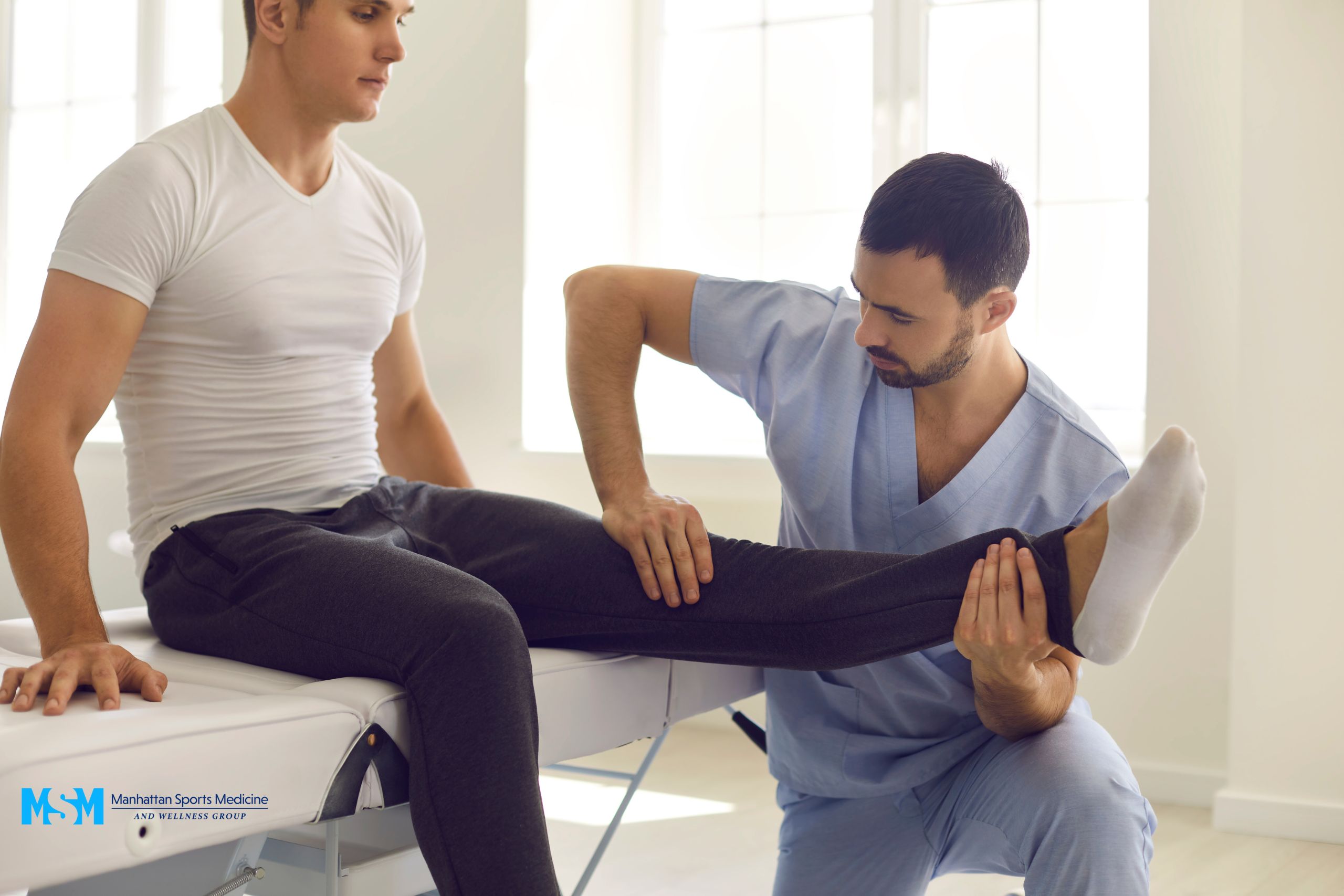 A physical therapist in blue scrubs assists a man seated on an examination table by stretching his leg. A physical therapist in blue scrubs assists a man seated on an examination table by stretching his leg