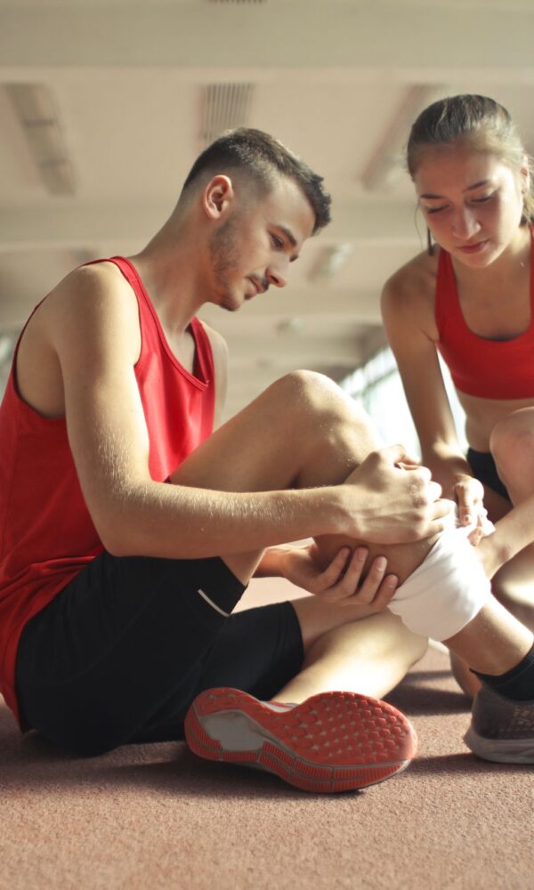 A woman helps a man with a wrapped ankle on an indoor track Both are wearing red athletic clothing