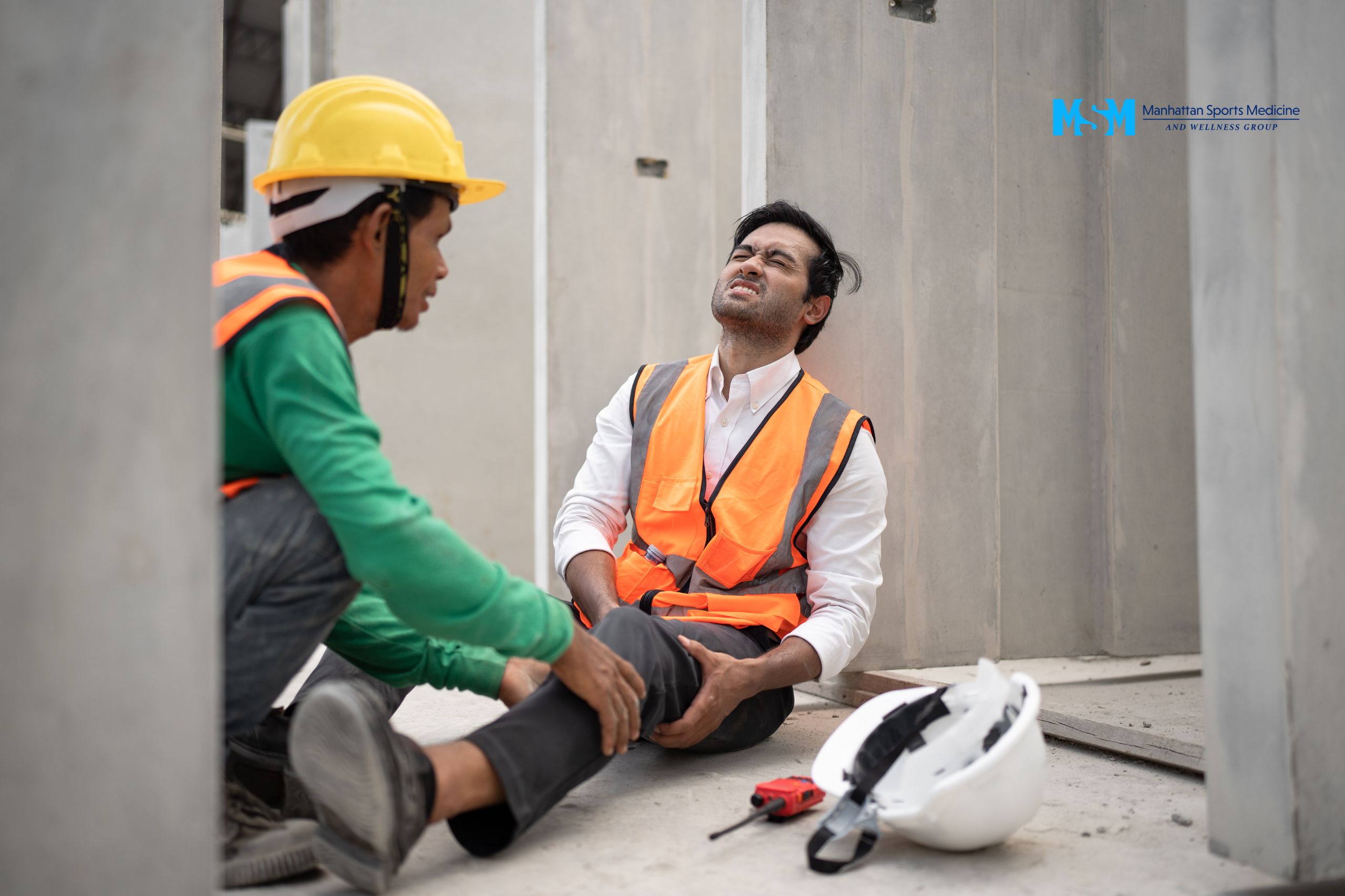A construction worker sits on the ground holding his knee, appearing in pain, while another worker crouches beside him wearing a yellow hard hat. A helmet and tools are on the floor. A construction worker sits on the ground holding his knee, appearing in pain, while another worker crouches beside him wearing a yellow hard hat A helmet and tools are on the floor