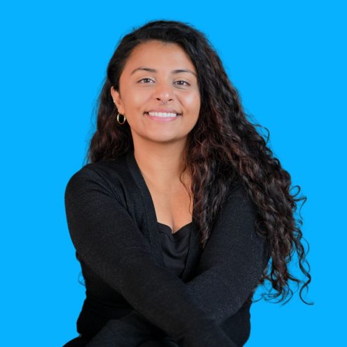 A woman with long curly hair wearing a black top is sitting with her arms crossed and smiling in front of a bright blue background. A woman with long curly hair wearing a black top is sitting with her arms crossed and smiling in front of a bright blue background.