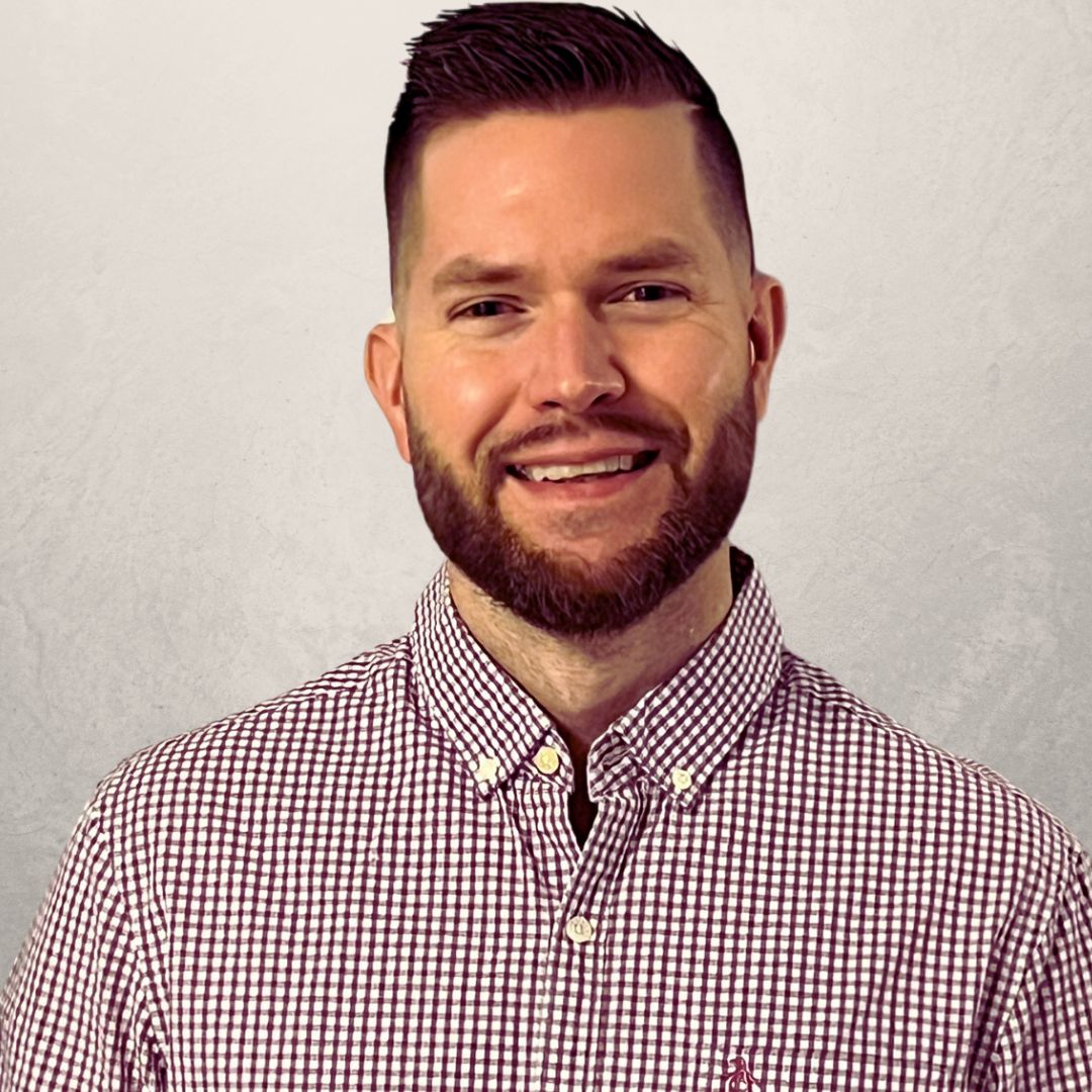 A man with short hair and a beard, wearing a red and white checkered button-up shirt, stands in front of a plain light-colored background, smiling at the camera. A man with short hair and a beard, wearing a red and white checkered button up shirt, stands in front of a plain light colored background, smiling at the camera