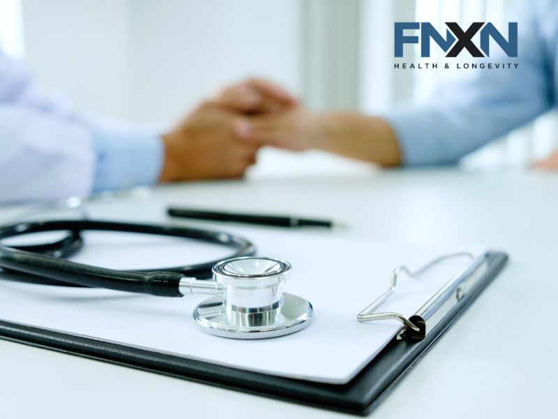 A stethoscope rests on a clipboard in the foreground while two people hold hands across a desk in the background, with a FNXN Health & Longevity logo. A stethoscope rests on a clipboard in the foreground while two people hold hands across a desk in the background, with a FNXN Health & Longevity logo