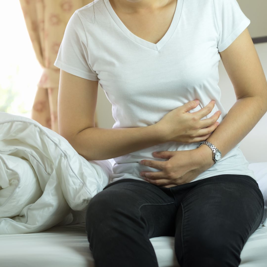 A person sitting on a bed holds their stomach with both hands, appearing to experience abdominal discomfort. A person sitting on a bed holds their stomach with both hands, appearing to experience abdominal discomfort